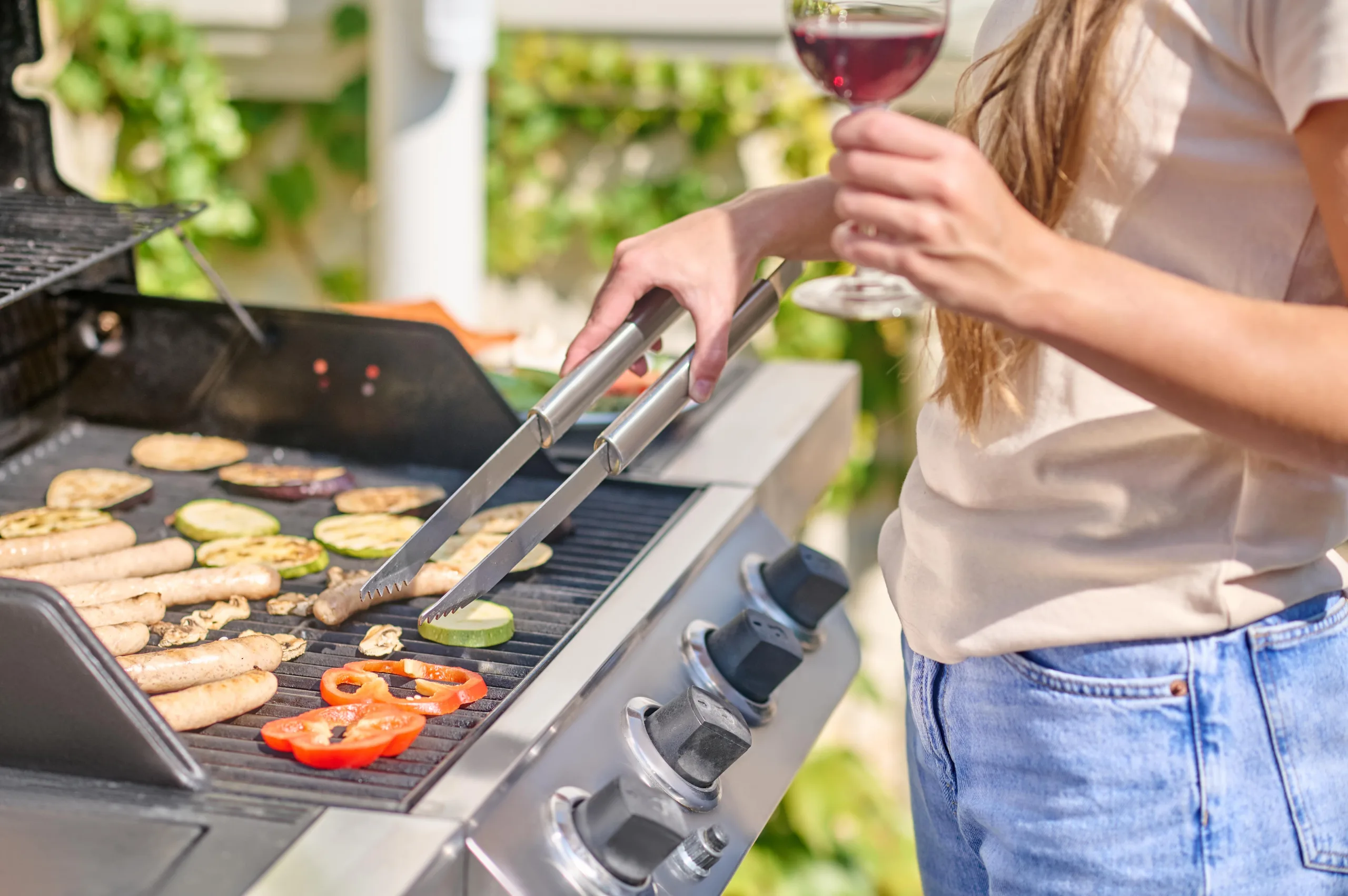Woman grilling turning sausages and vegetables over with metal tongs. She is holding a glass of red wine, there is green foliage behind her, and she is wearing a loose t-shirt and denim jeans.