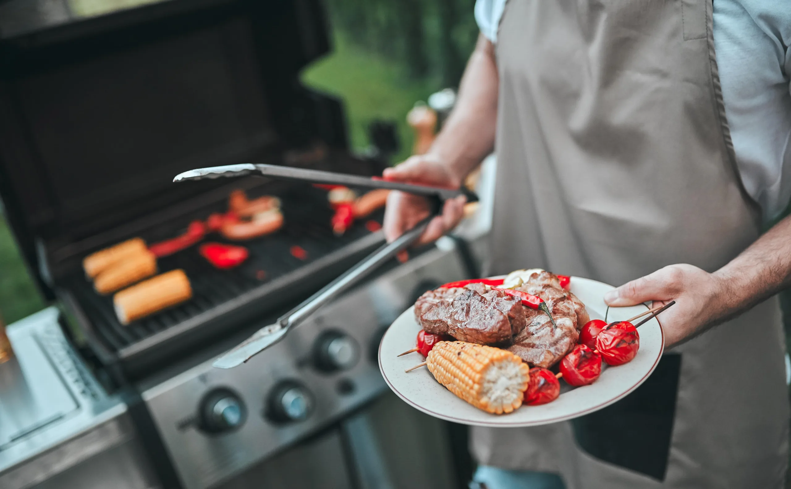Man holding a plate in front of a grill. The plate has freshly grilled corn, kabobs, and steak and chili peppers. Her is wearing an apron and tongs seeming like he is about to hand the plate to someone before returning to tend to the grill.